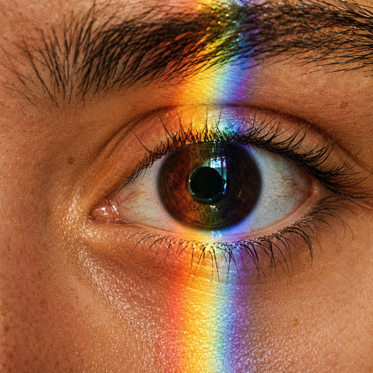 Extreme Nahaufnahme eines menschlichen Auges mit einem vertikalen Regenbogen-Lichtreflex über Augenlid, Iris und Haut.