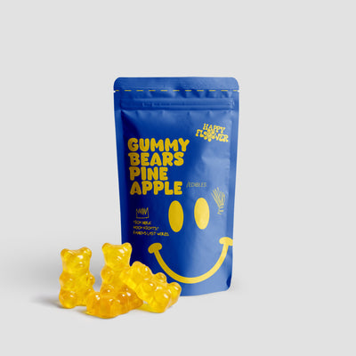 Happy Flower Gummy Bears Pineapple Edibles in blauer Standbeutel-Verpackung mit mehreren gelben Gummibärchen im Vordergrund auf hellem Hintergrund.