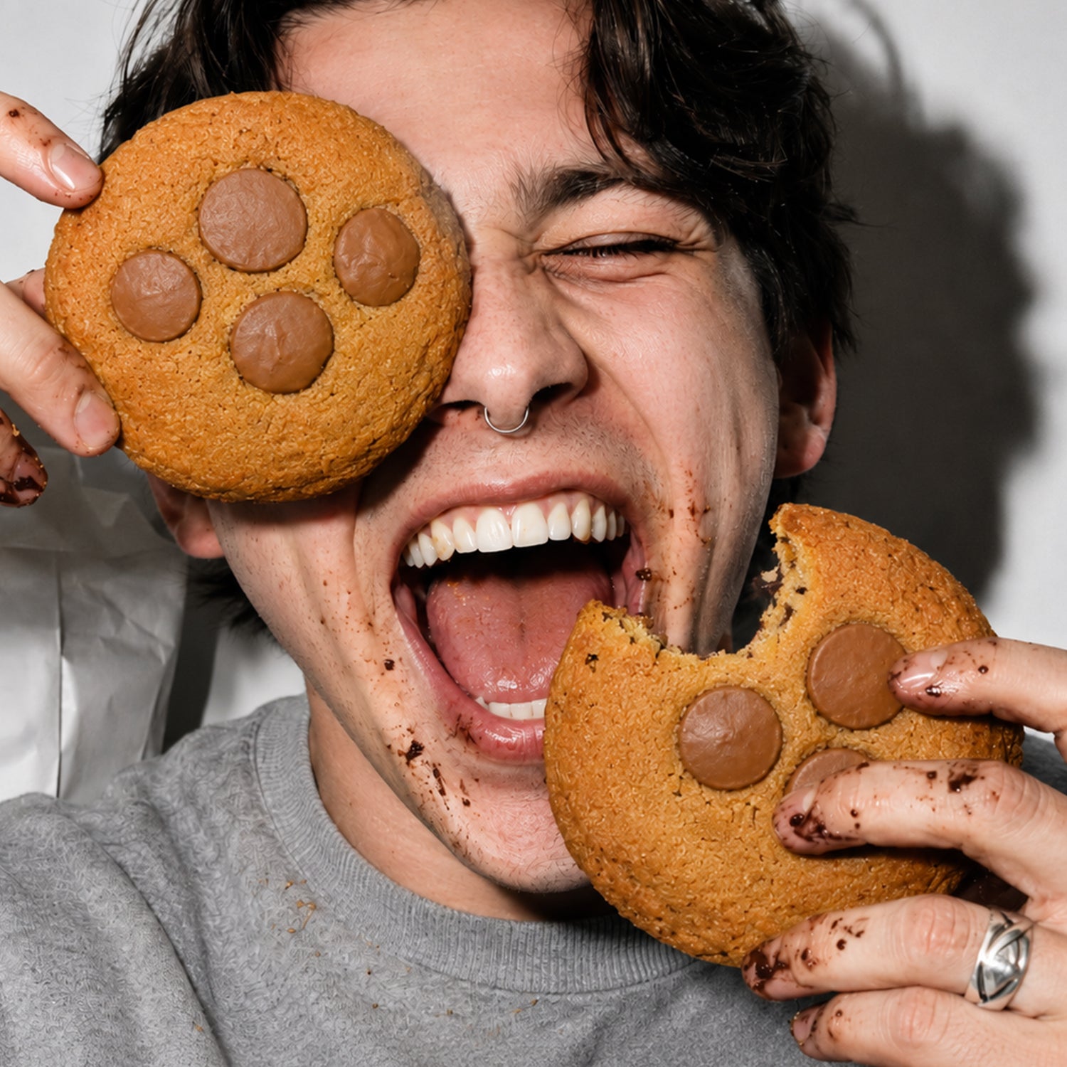 A smiling young man with dark hair and a septum ring is holding two large THC chocolate cookies in front of his face, one of them like an eye patch. Chocolate crumbs can be seen on his face and hands.