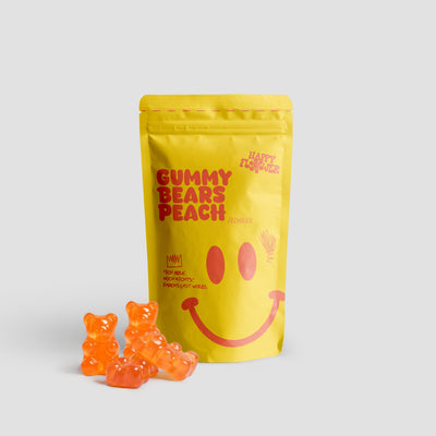Happy Flower Gummy Bears Peach Edibles in gelber Standbeutel-Verpackung mit mehreren orangefarbenen Gummibärchen im Vordergrund auf hellem Hintergrund.